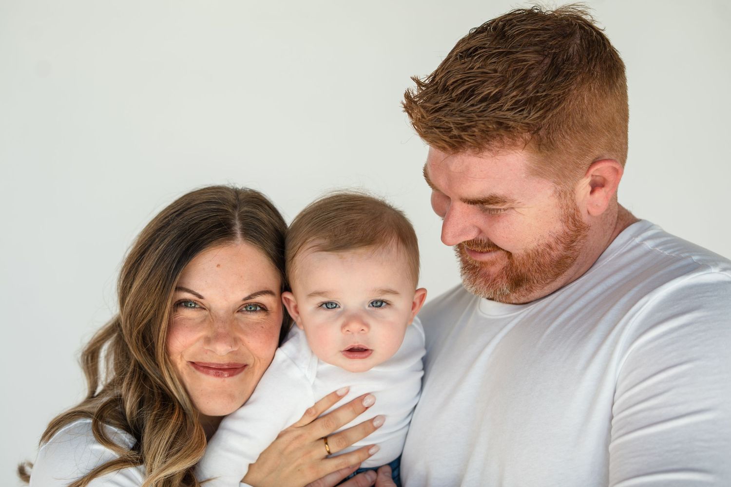 A smiling couple in white tops holds their adorable blue-eyed baby against a clean white background.