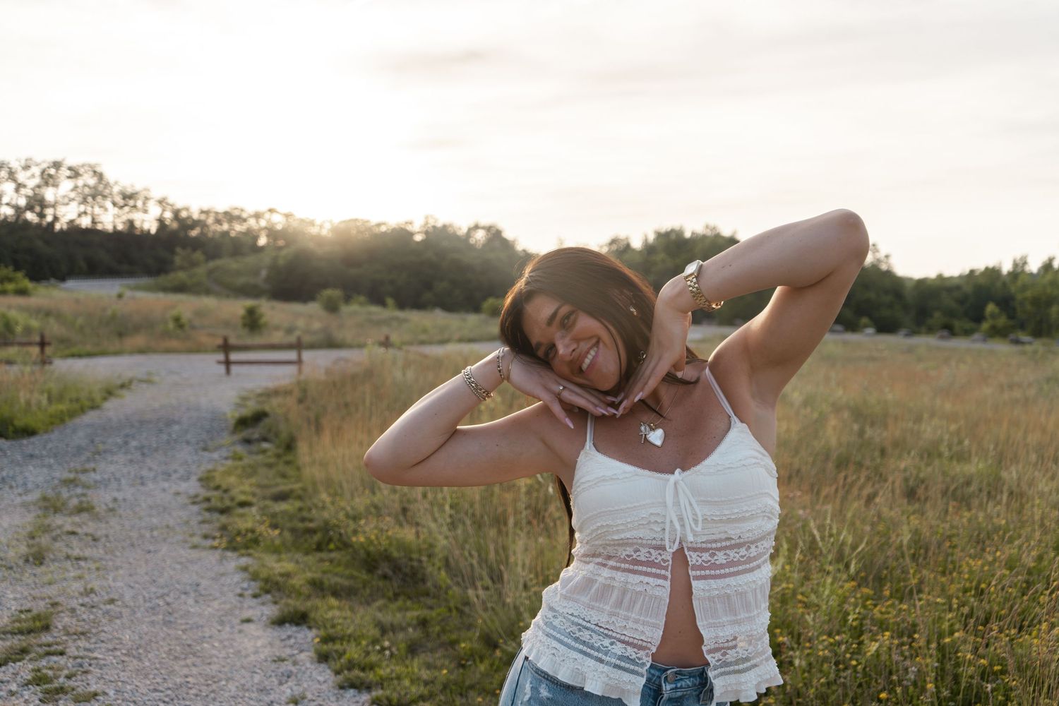 Person in white tank top poses on a gravel path during golden hour in a summer meadow.