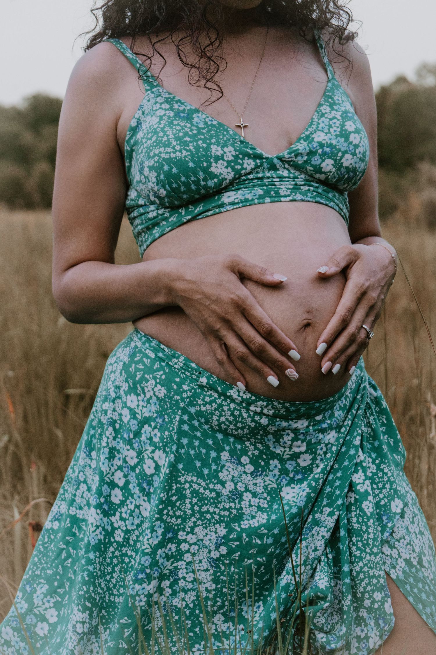 A person wearing a turquoise floral dress holds their stomach while standing in a field of tall grass at sunset.
