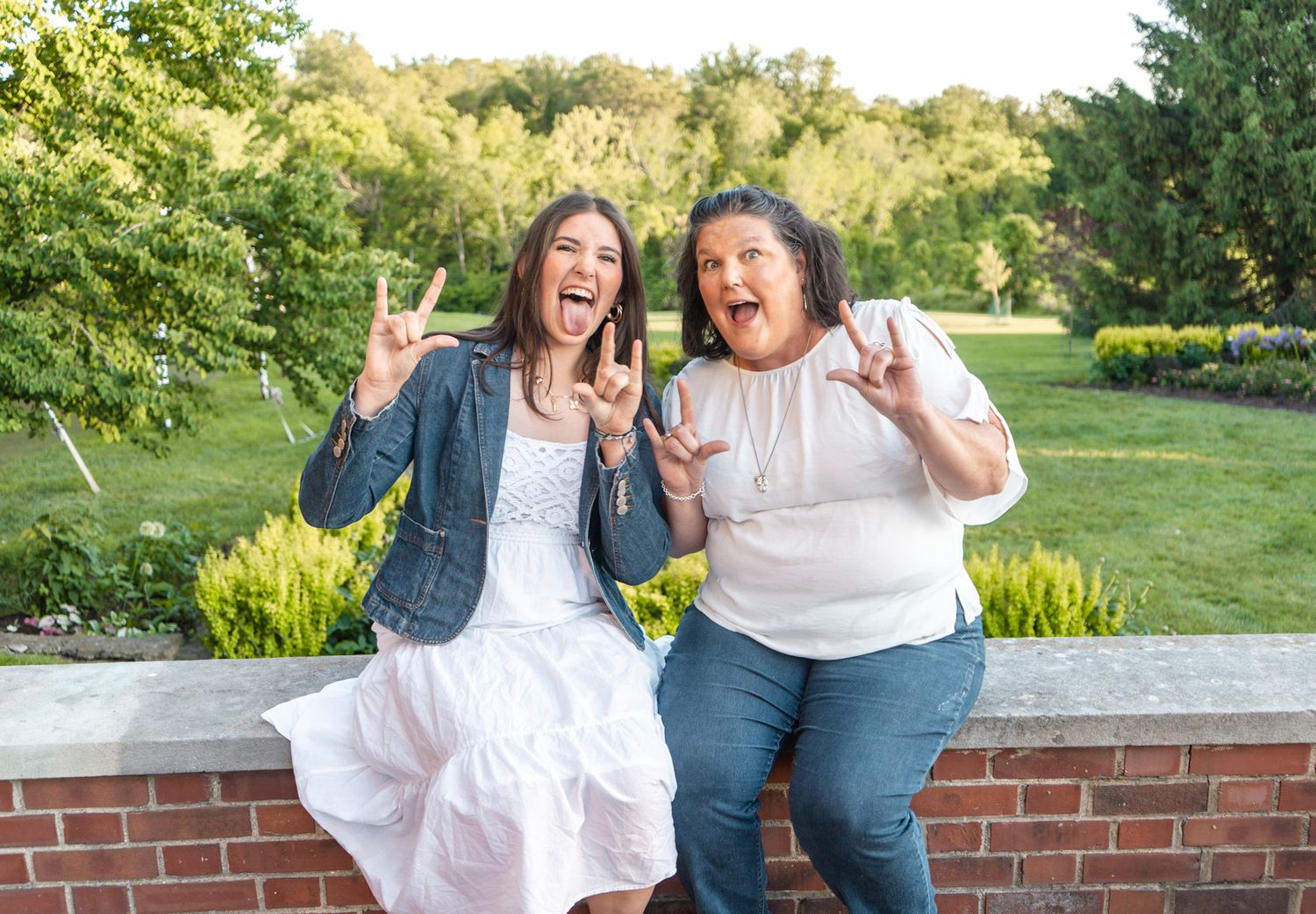 Two friends sitting on a brick wall outdoors sharing a joyful moment with peace signs and laughter.