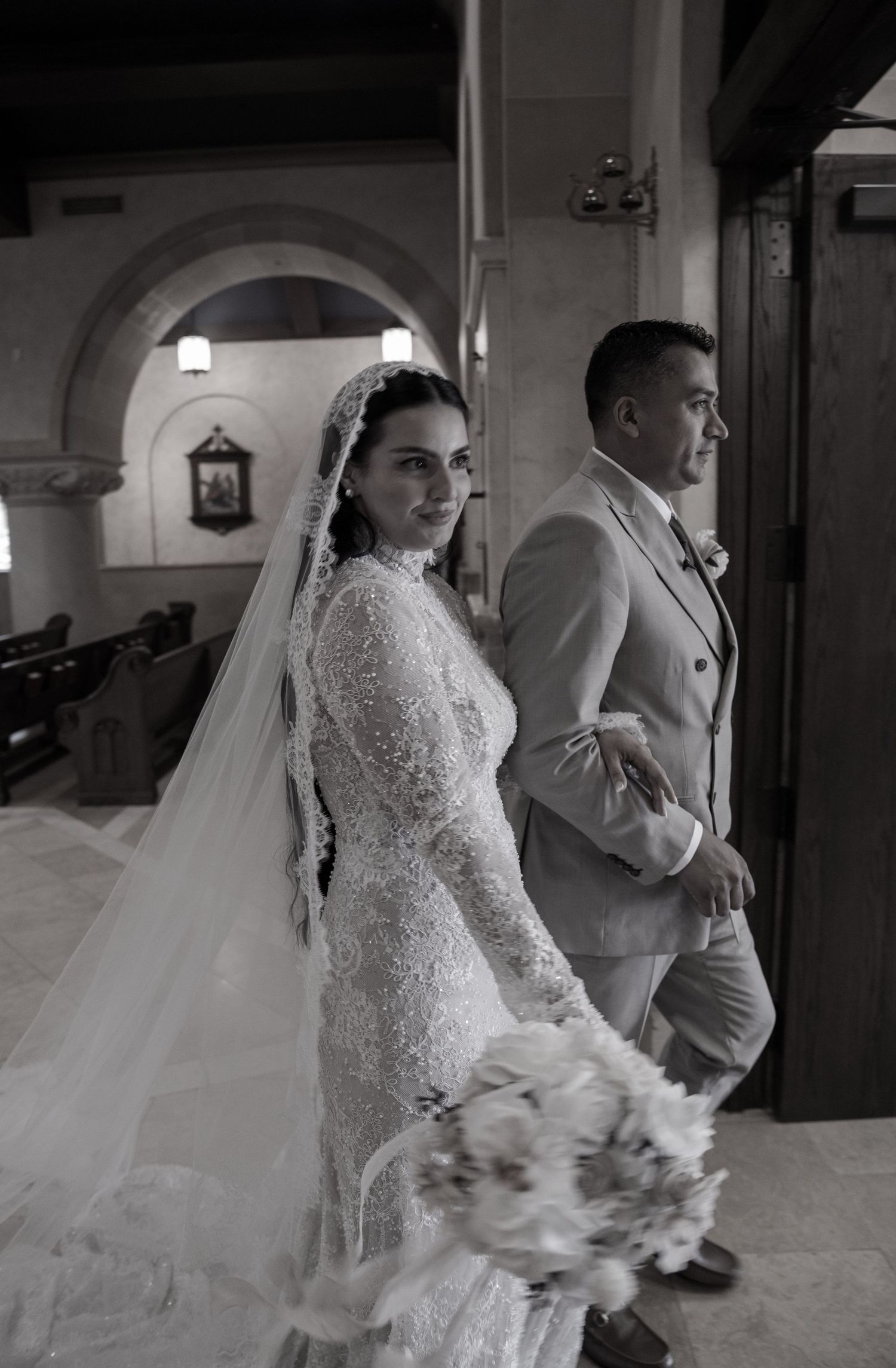 Black and white photo of a bride in lace gown and veil holding bouquet beside groom in suit inside a church.