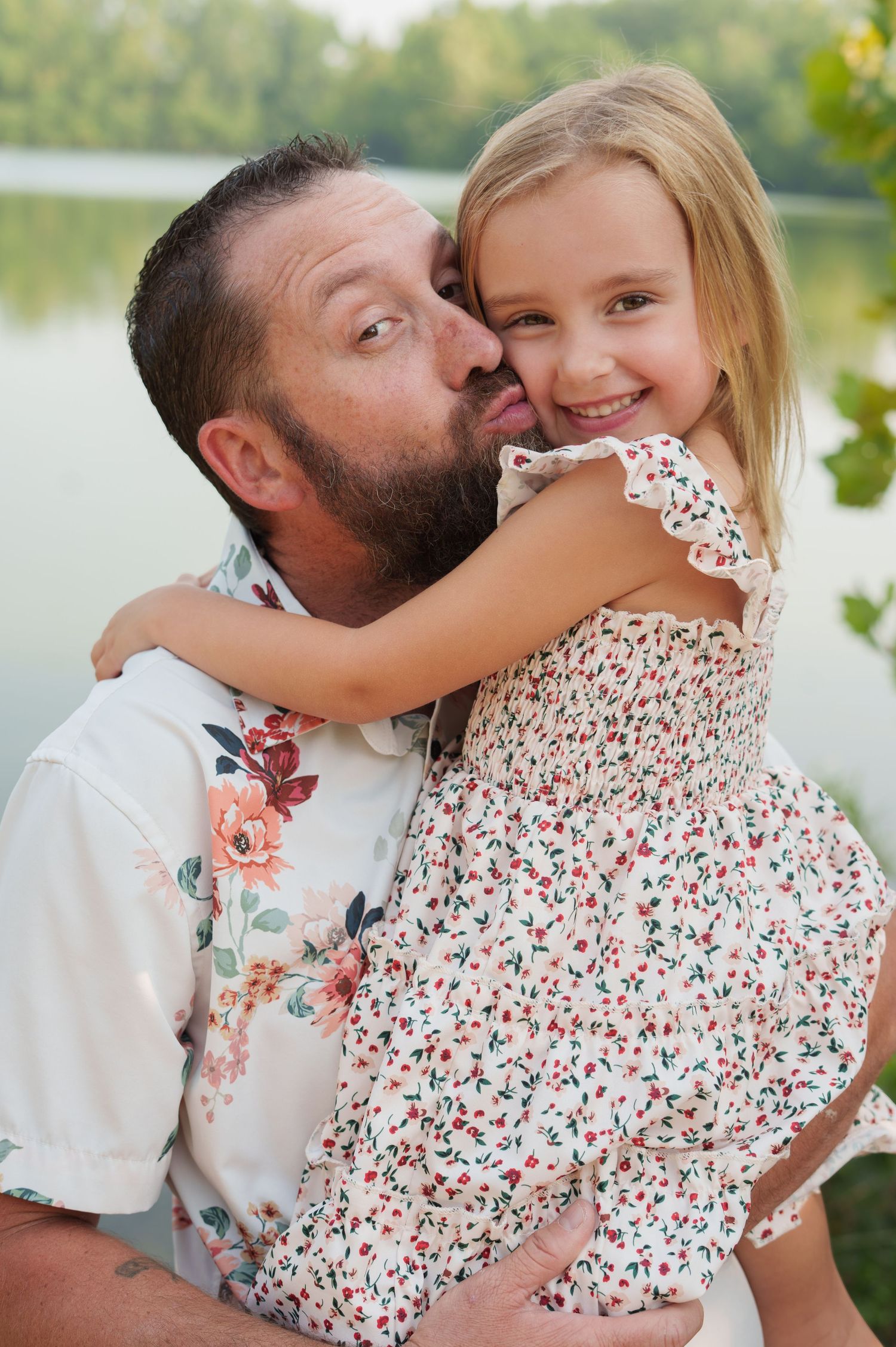 A bearded man in a floral shirt kisses a smiling young girl in a matching floral dress outdoors near a lake.
