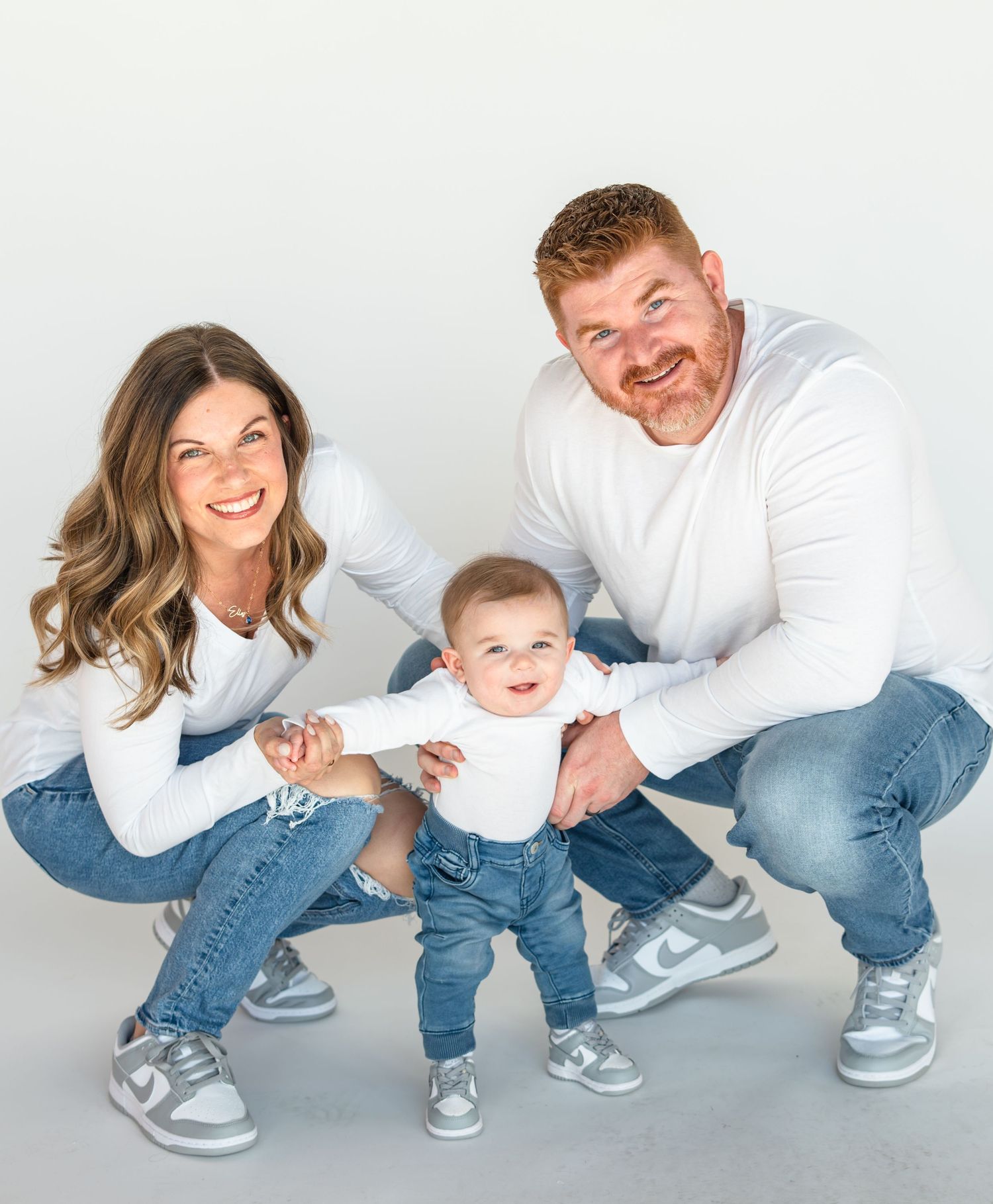 A happy family of three poses in matching white shirts and jeans, with parents holding their smiling baby upright.