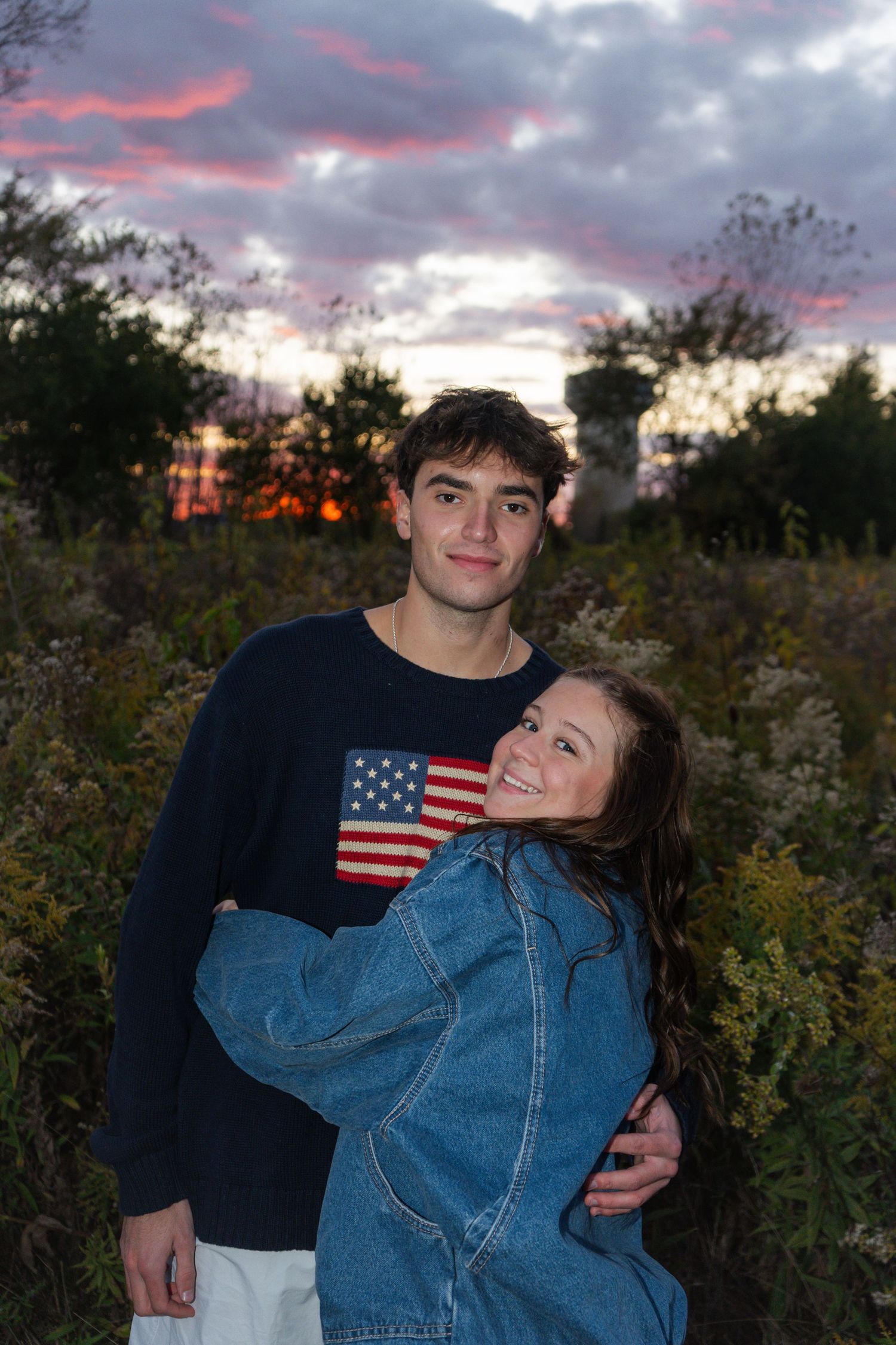 A couple embraces in an outdoor setting with American flag sweater and denim jacket at dusk.