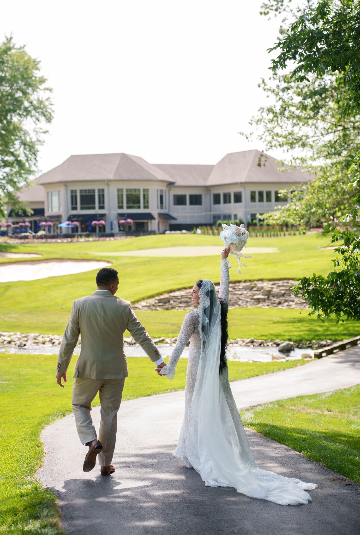 Bride and groom walking hand in hand on a path toward a golf club venue, bride raising bouquet in celebration.