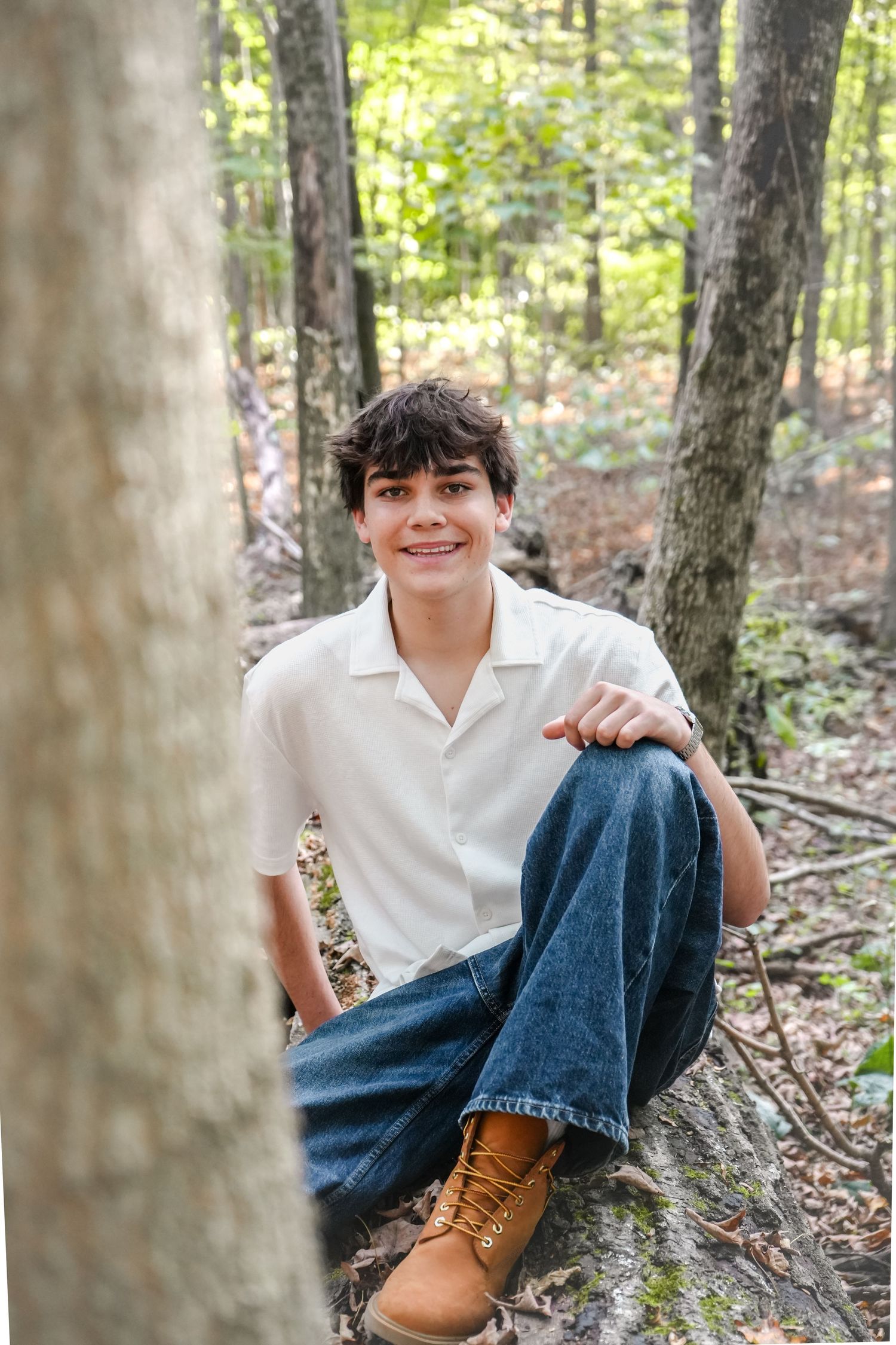 Smiling teenage boy sitting on forest floor wearing white shirt, jeans, and tan boots among tall trees.