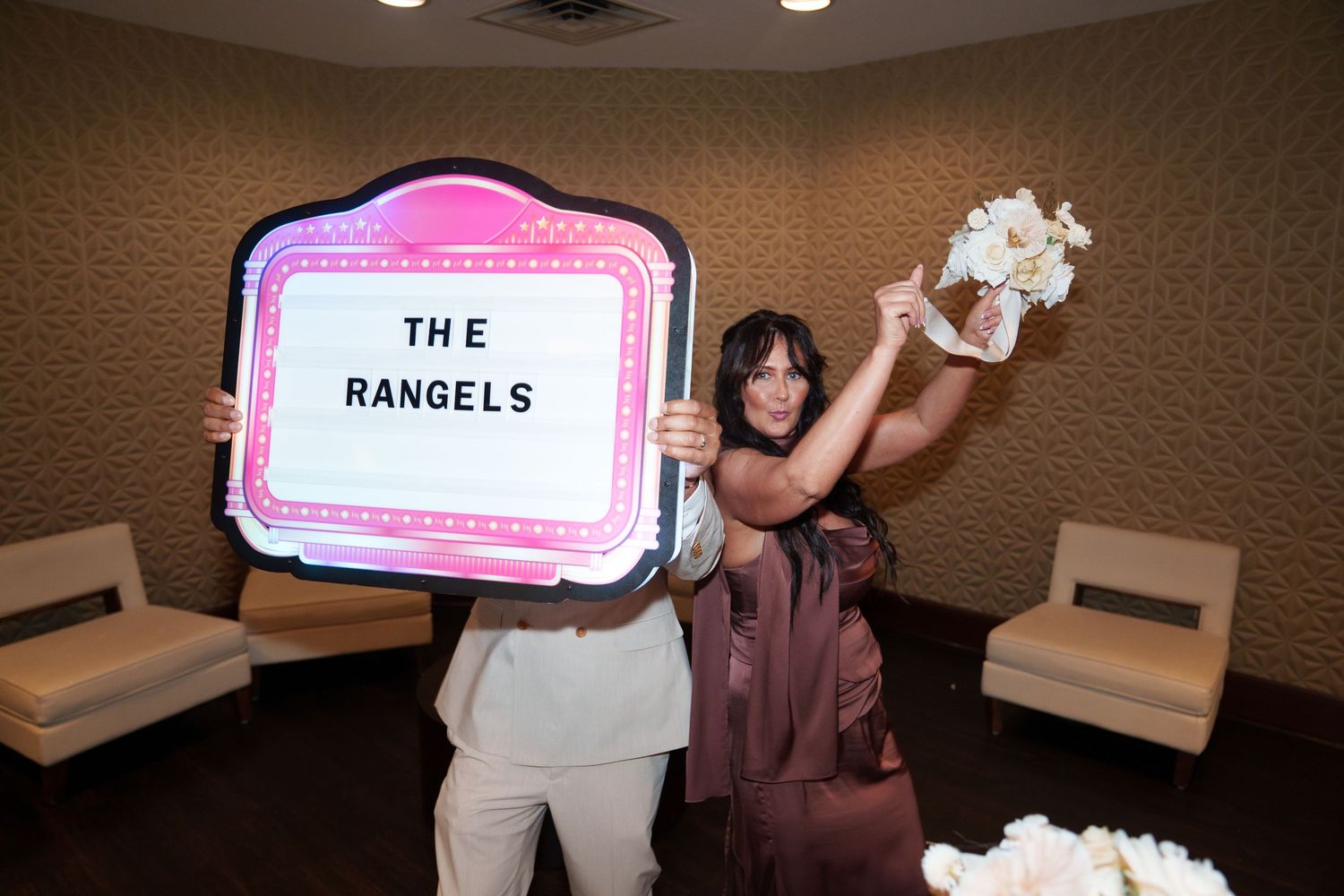 Two women celebrate at a wedding reception holding a decorative sign reading The Rangels and a bridal bouquet.