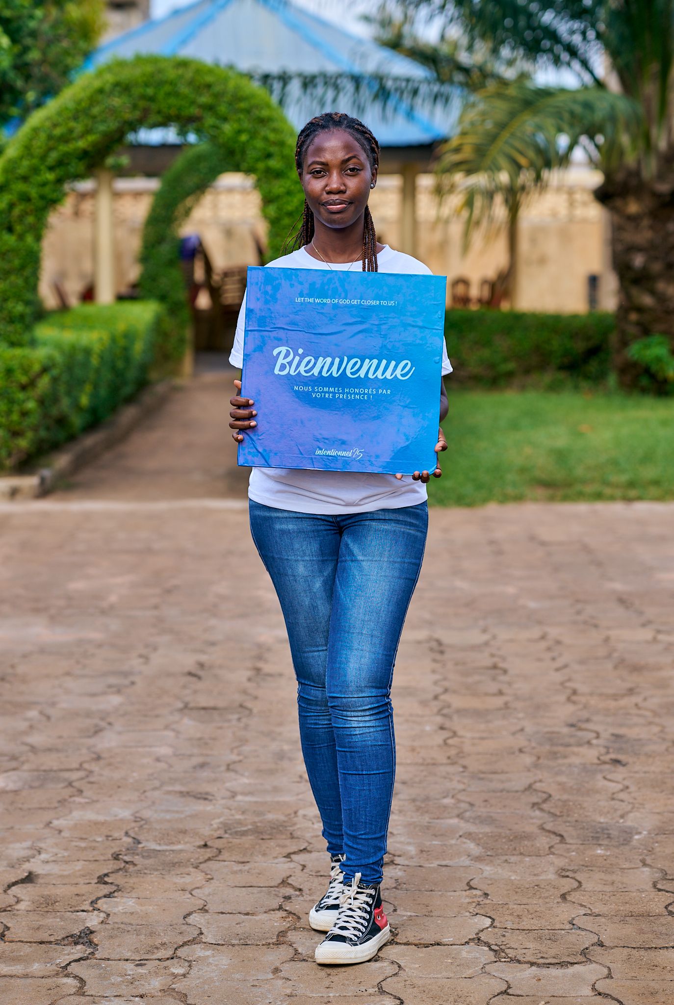 Person in jeans holding blue welcome sign on brick pathway surrounded by gardens at sharing moment called intentionnel