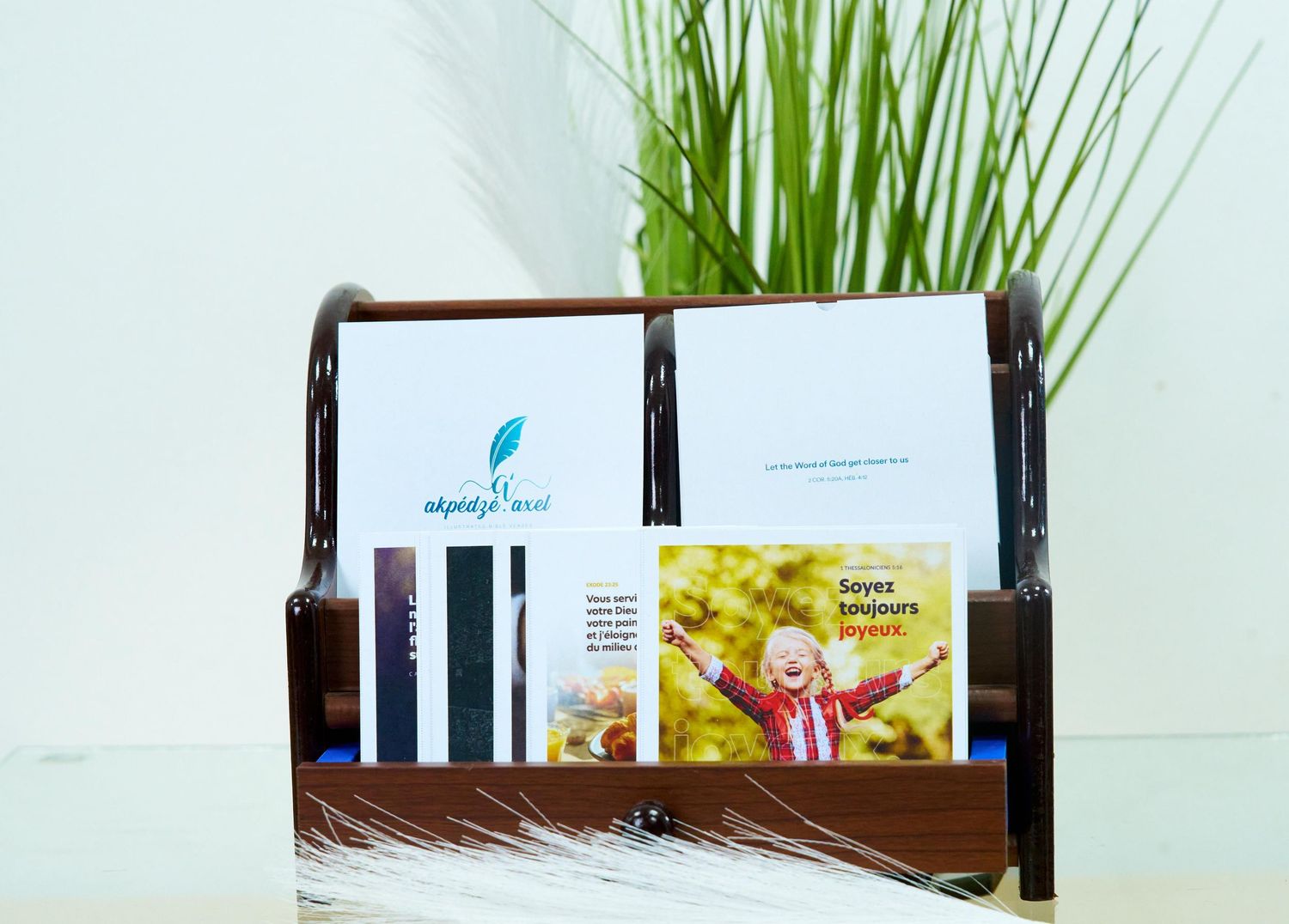Marketing materials arranged in dark wood display stand with decorative grass plant. Verset biblique illustré, illustrated biblical verse