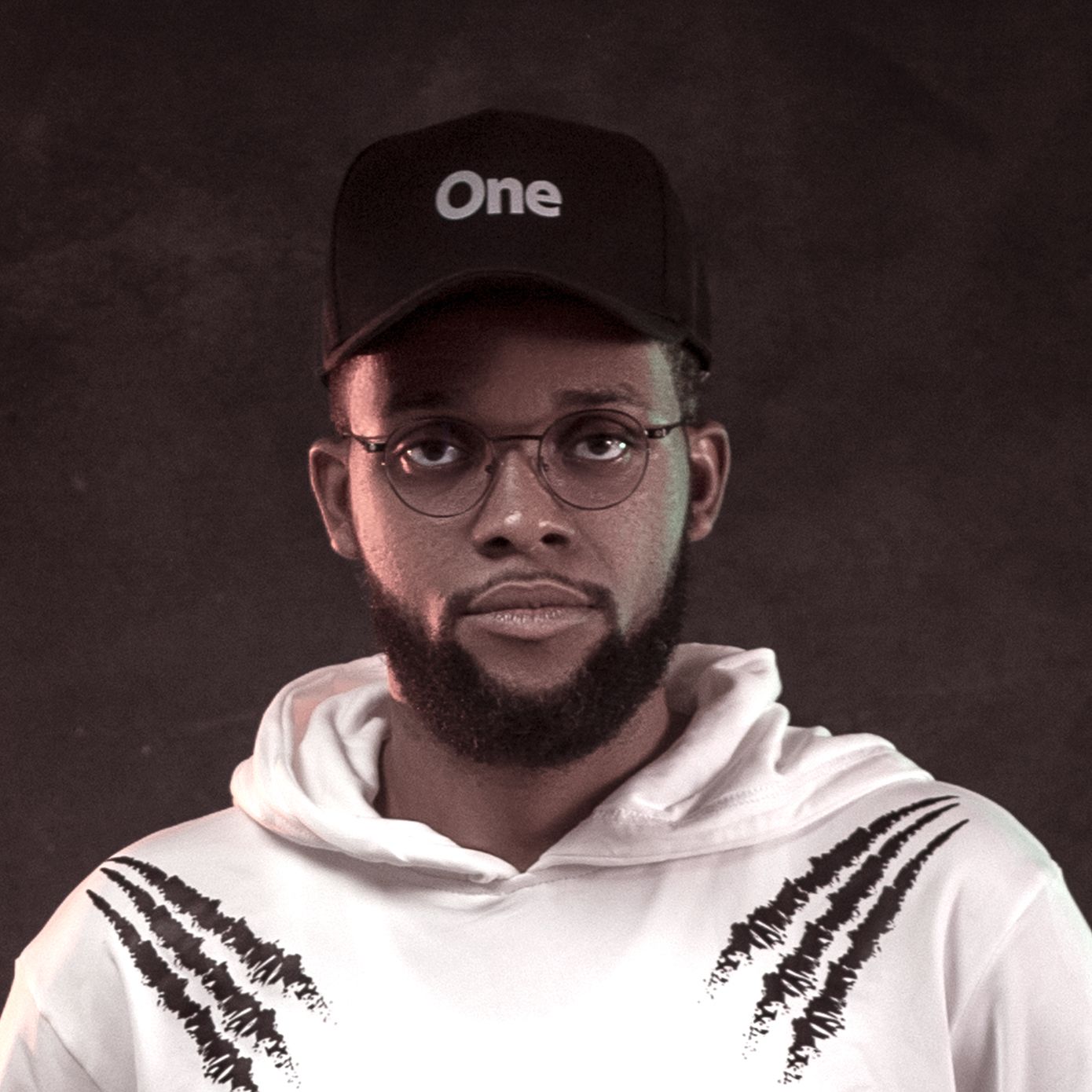 Portrait of person in white hoodie and black cap with 'One' text against dark background.
