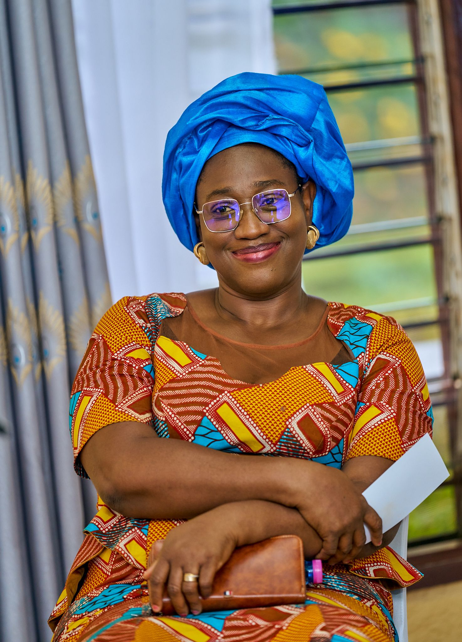 Woman wearing colorful African print dress and bright blue head coverings smile while seated at sharing moment called intentionnel
