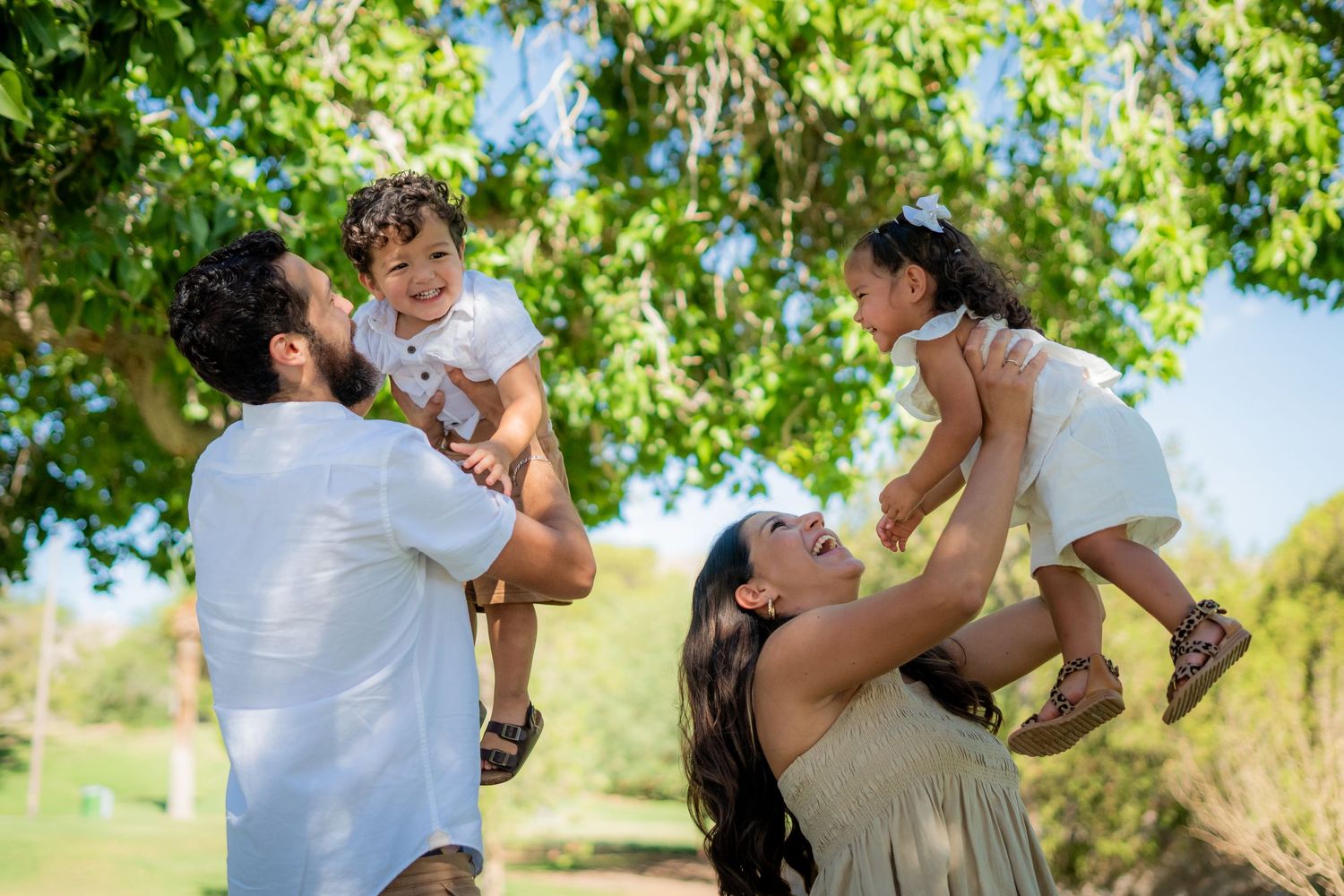 Family of four enjoying a sunny day in the park, parents lifting two toddlers up in the air under green trees.