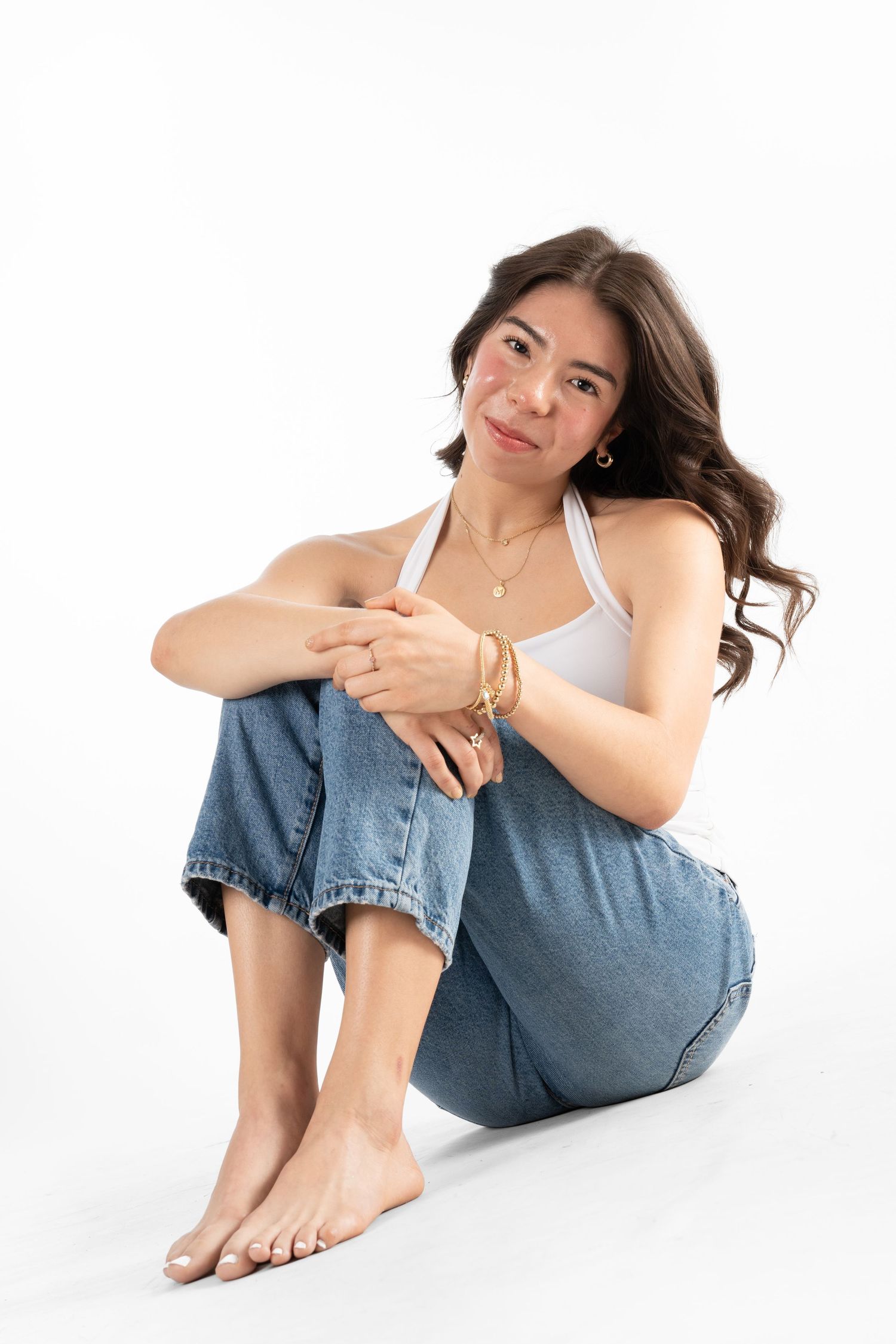 Young woman sitting on white background wearing white tank top, blue jeans, gold jewelry, with wavy brown hair.