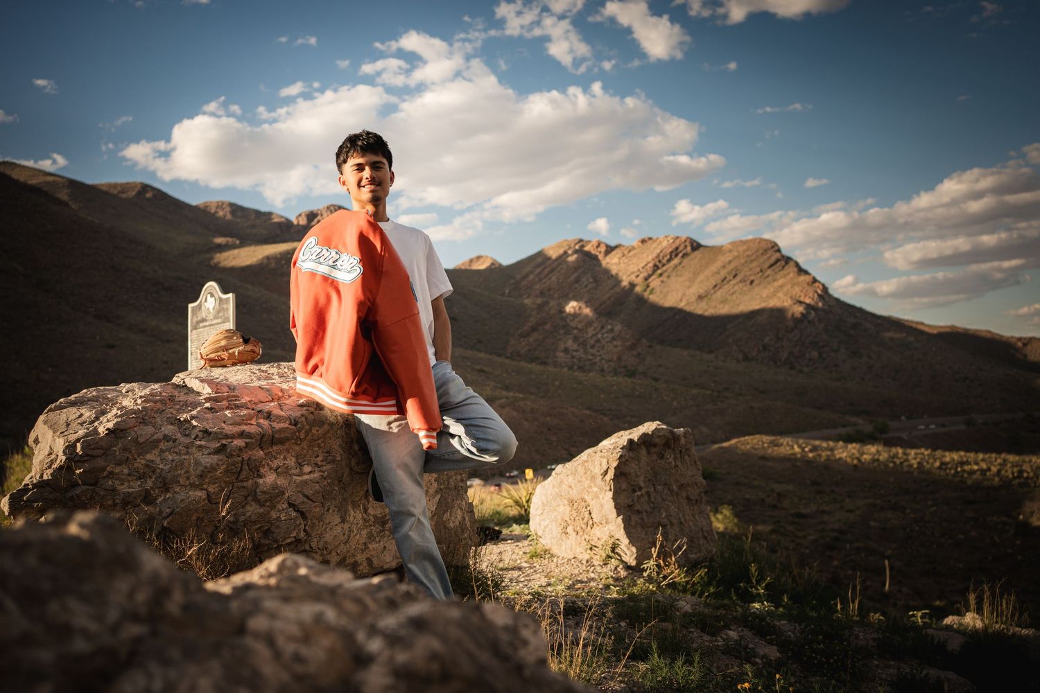 Young man sitting on rocks with scenic mountain landscape and dramatic cloudy sky in background.