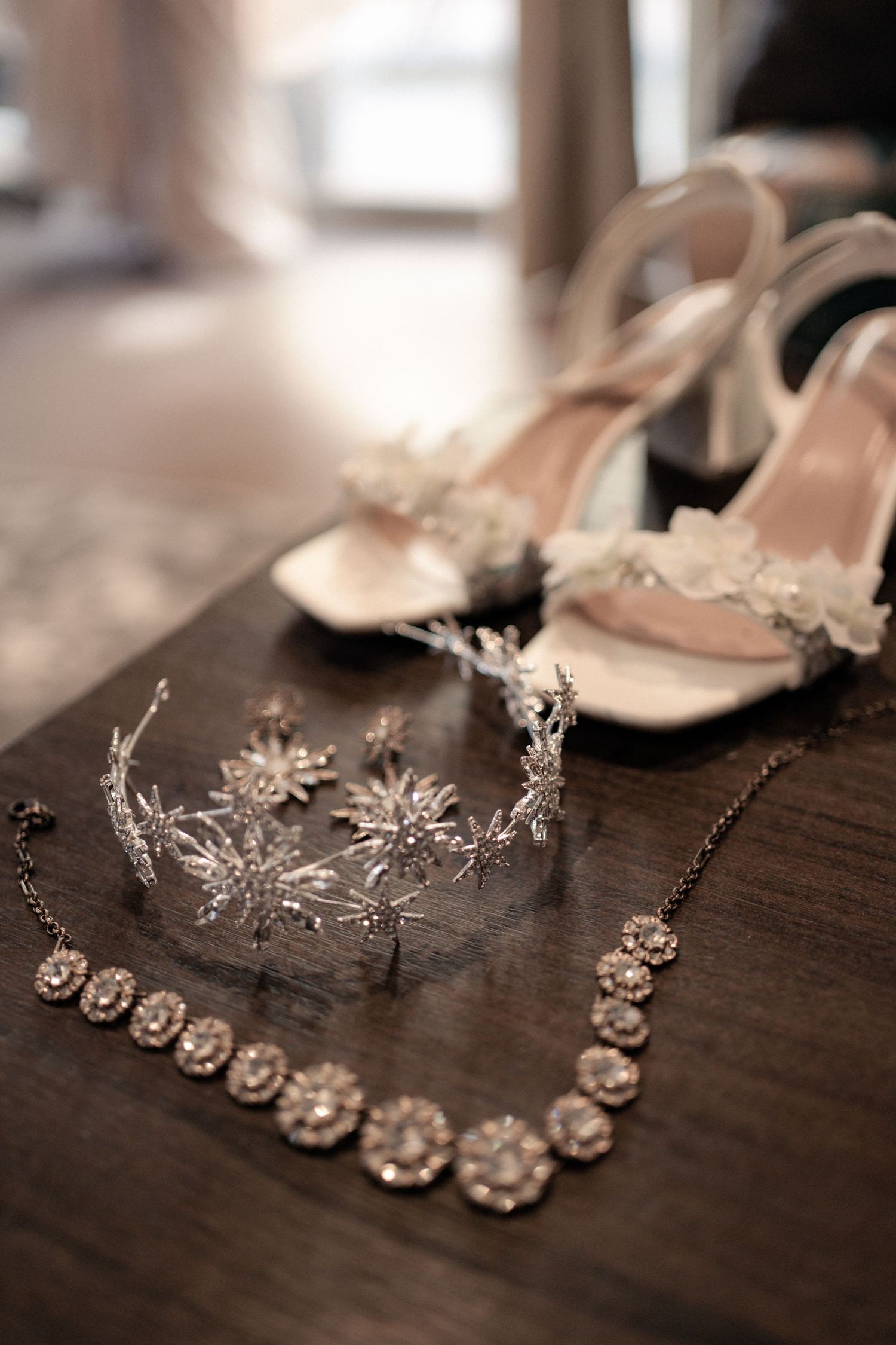 Elegant wedding accessories including peach heels, delicate silver tiara, and crystal floral necklace on dark wooden surface.