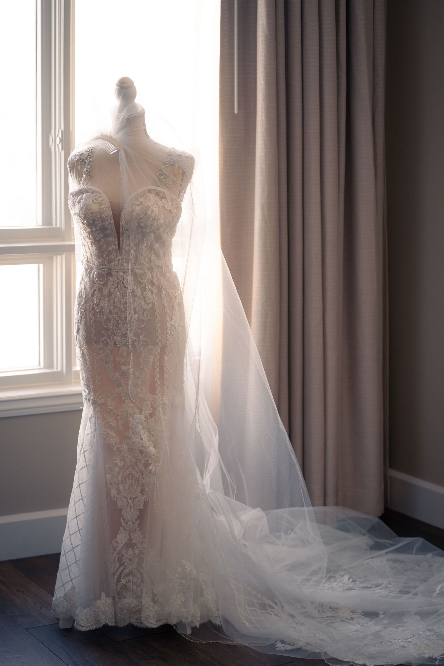 Elegant lace wedding dress displayed near window with flowing veil and natural lighting.