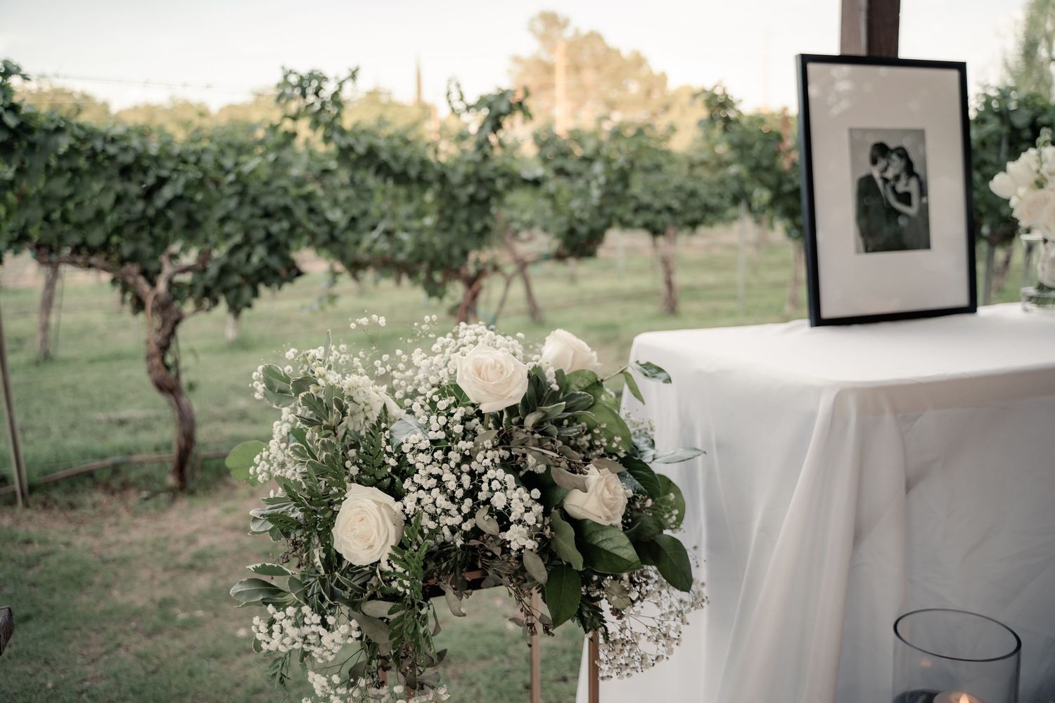 White floral arrangement with baby's breath sits beside a framed photo at an outdoor vineyard wedding reception.