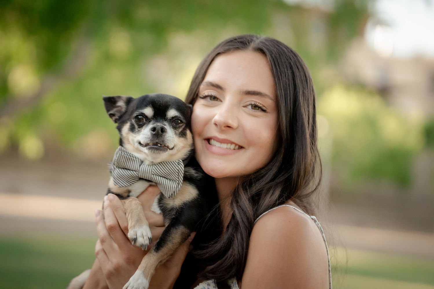 Smiling young woman holding a Chihuahua dog wearing a bow tie outdoors in a park setting.