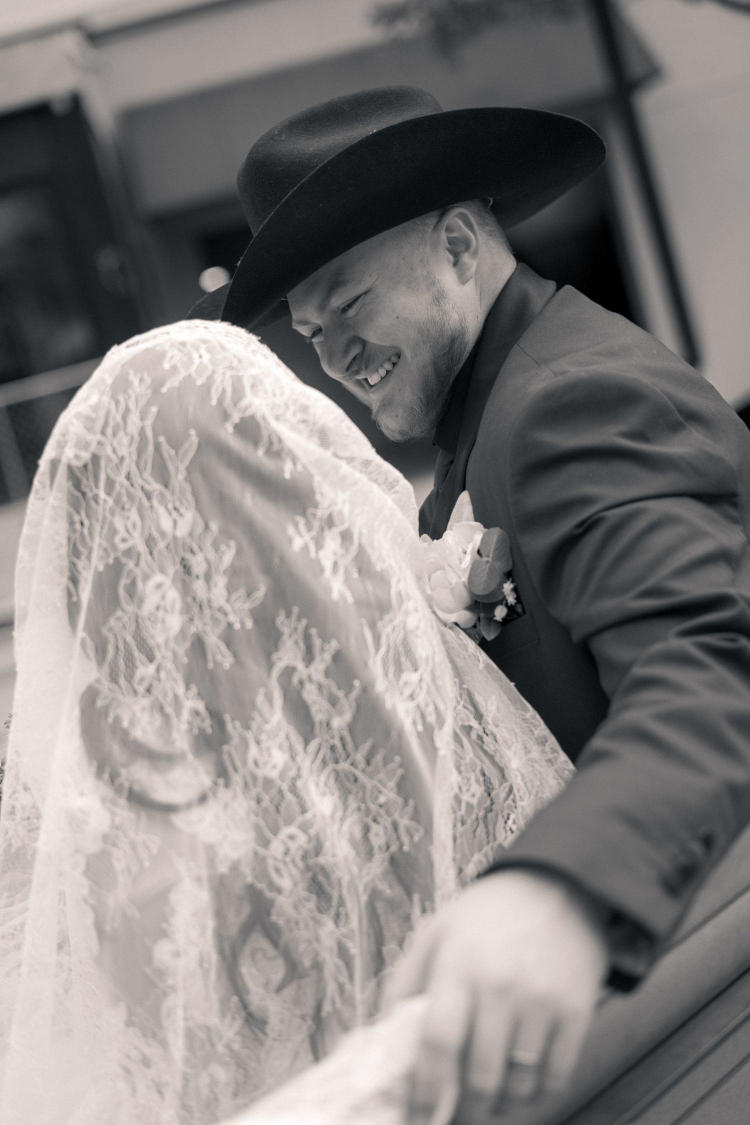 Black and white photo of a smiling groom in a cowboy hat leaning toward his bride in a lace veil at their wedding.