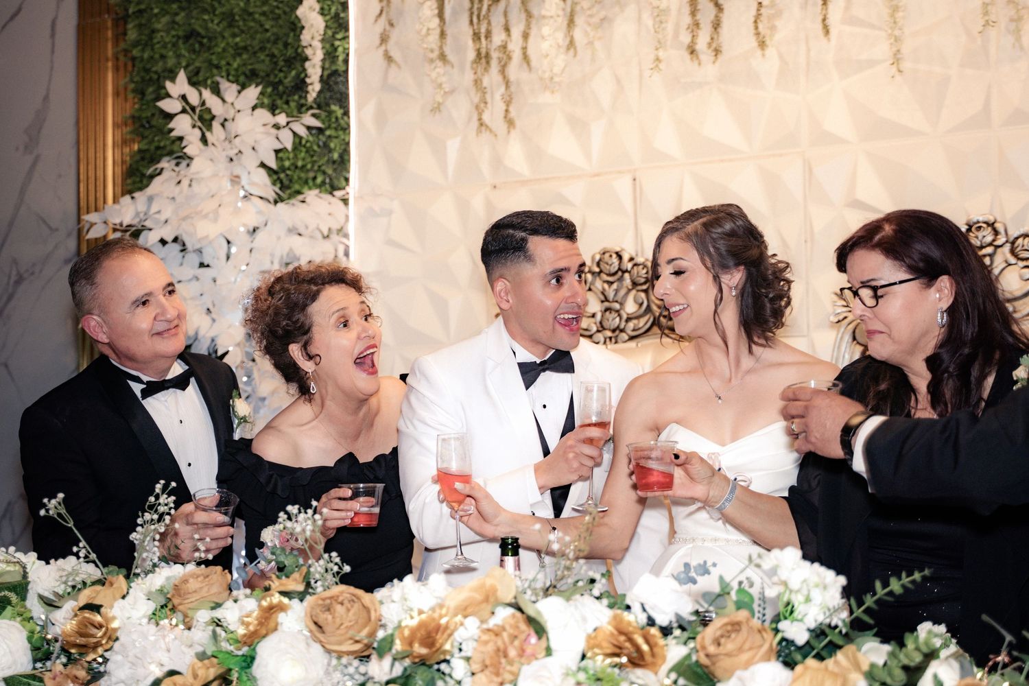 Bride and groom toast with family at elegant wedding reception with white floral arrangements and gold accents.