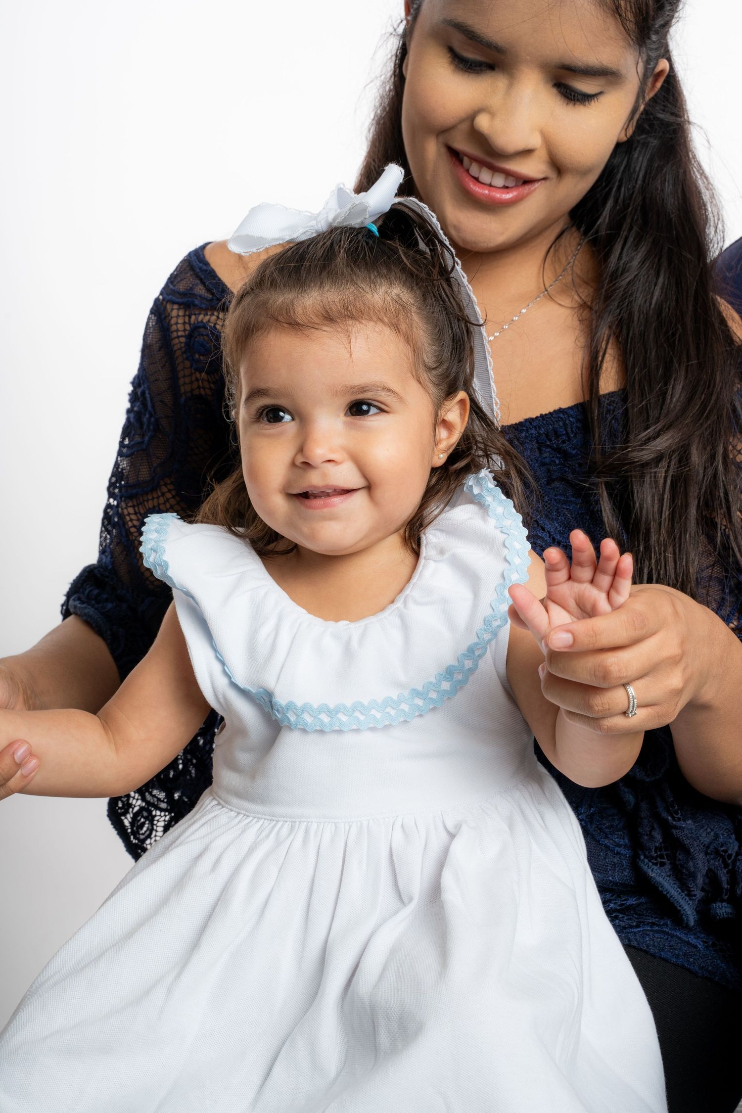 A smiling toddler girl in a white dress sits on a young woman's lap against a white background.