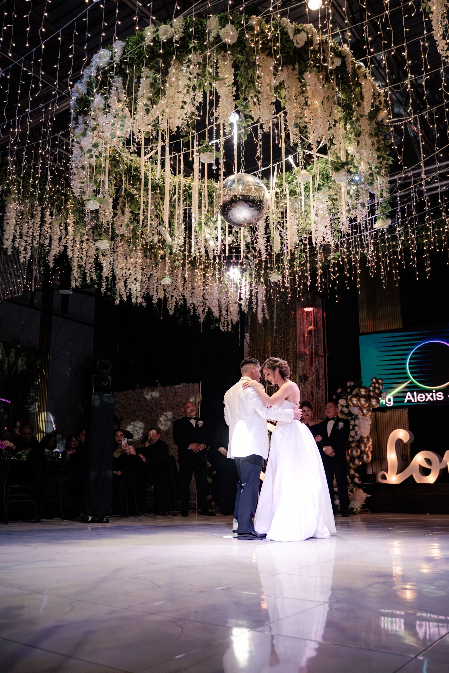 Bride and groom share first dance under floral chandelier and twinkling lights at elegant wedding reception.