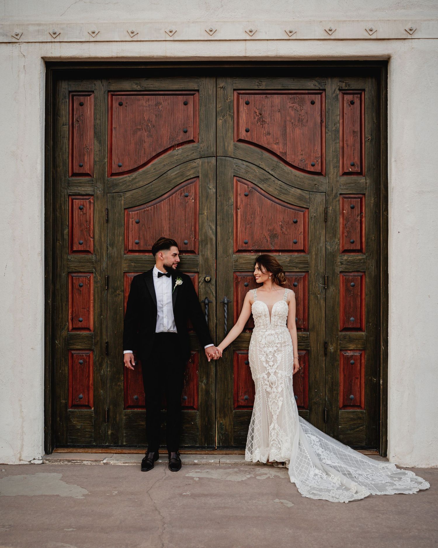 Bride in lace gown and groom in black tuxedo hold hands before ornate wooden doors on their wedding day.
