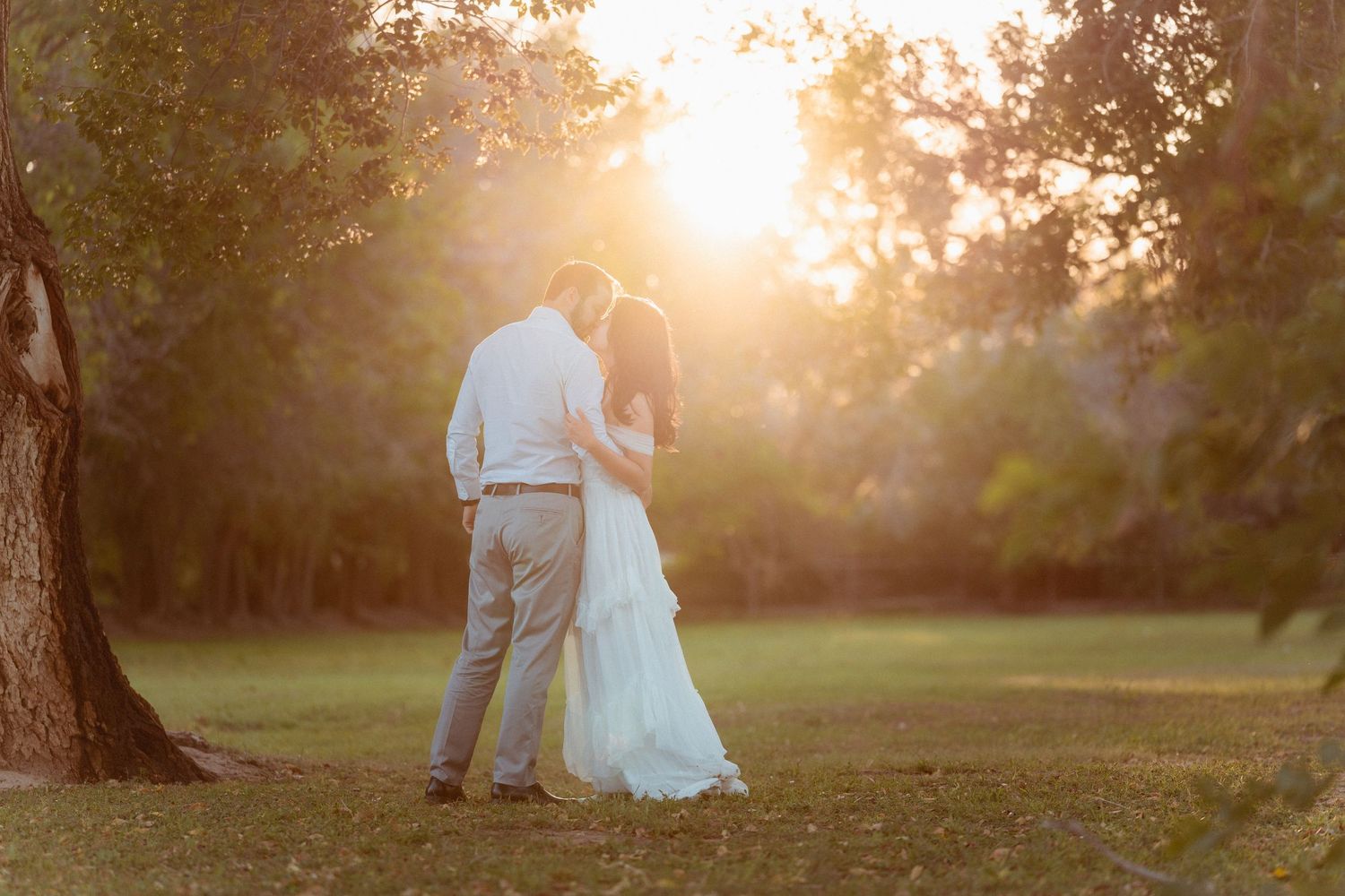 A couple embraces in a romantic sunset moment on a grassy lawn with golden sunlight filtering through trees.