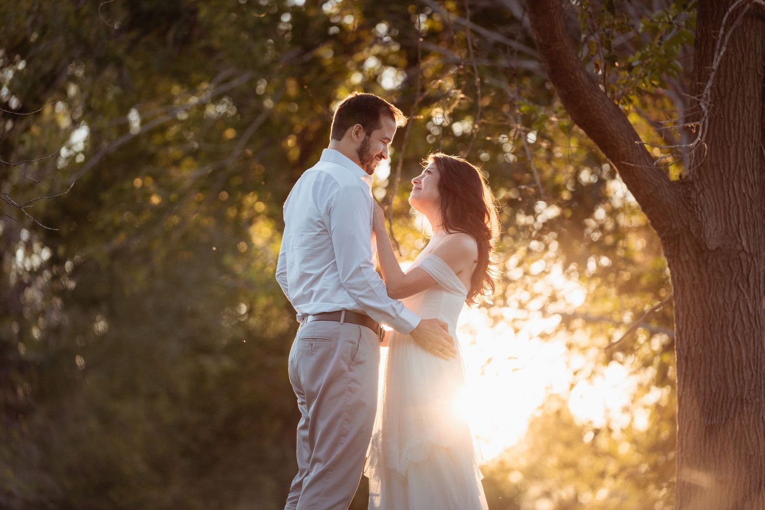 A couple embraces in a romantic outdoor setting with sunlight streaming through trees during golden hour.
