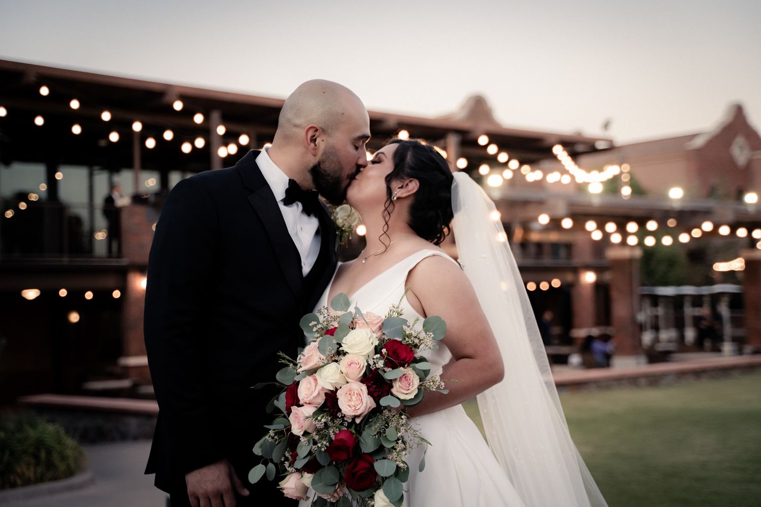 Romantic outdoor evening wedding portrait under string lights with pink floral bouquet.