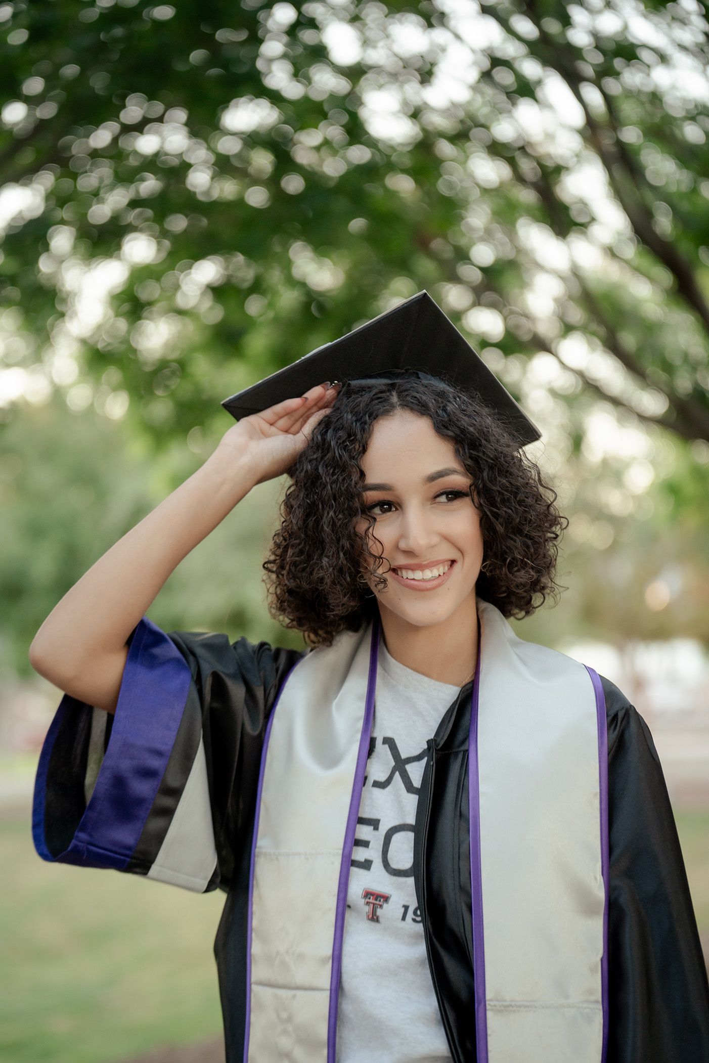 A graduate in academic regalia poses playfully in an outdoor setting with natural bokeh background.