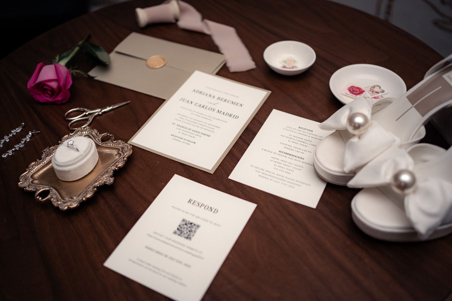 Wedding details including invitations, white shoes, and a pink rose arranged on a dark wooden table.