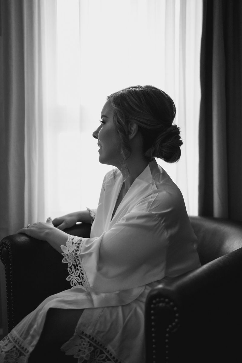 A bride in a white robe gazes thoughtfully out a window in an elegant black and white portrait.