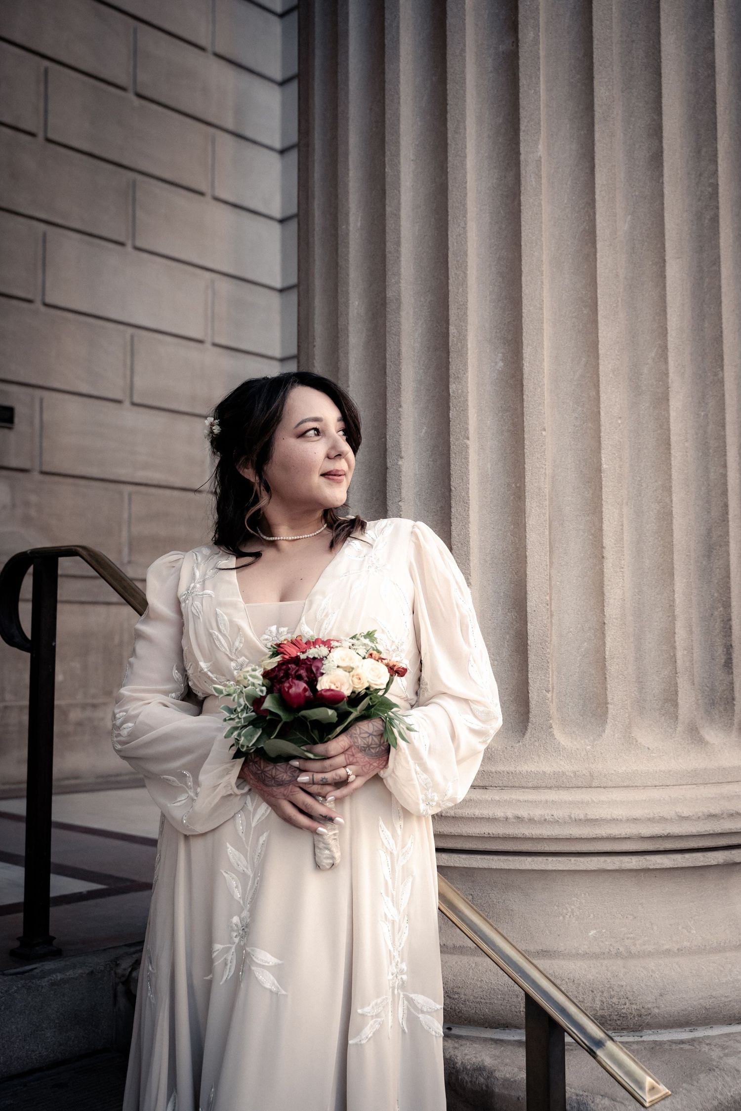 Bride in vintage-style dress holds bouquet while standing by classical architecture.