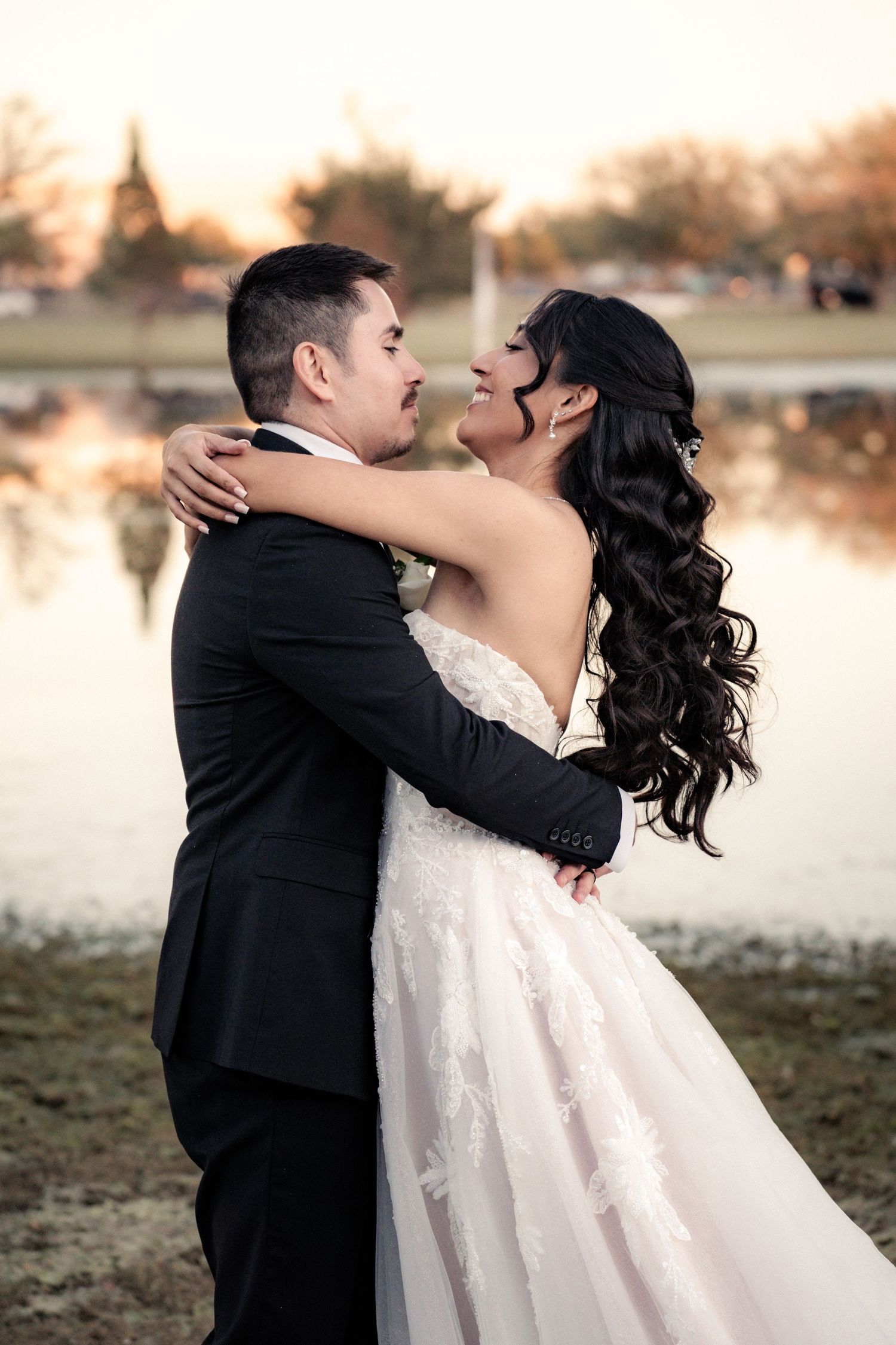 Bride and groom embrace at sunset near a reflective pond, bride in lace gown with long curly hair, groom in black tuxedo.