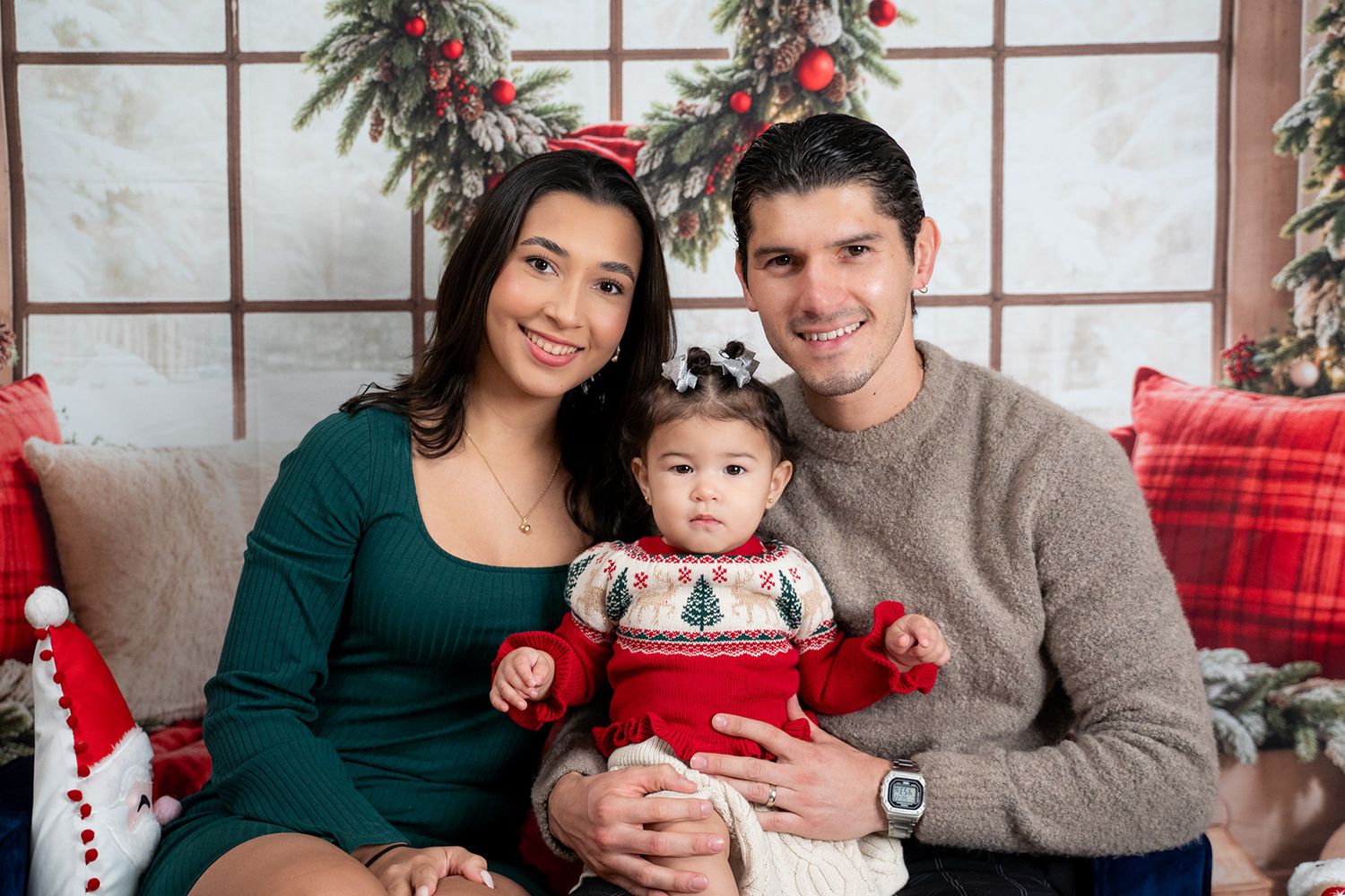 A family wearing green and grey sweaters poses together for holiday photos with festive red decorations.