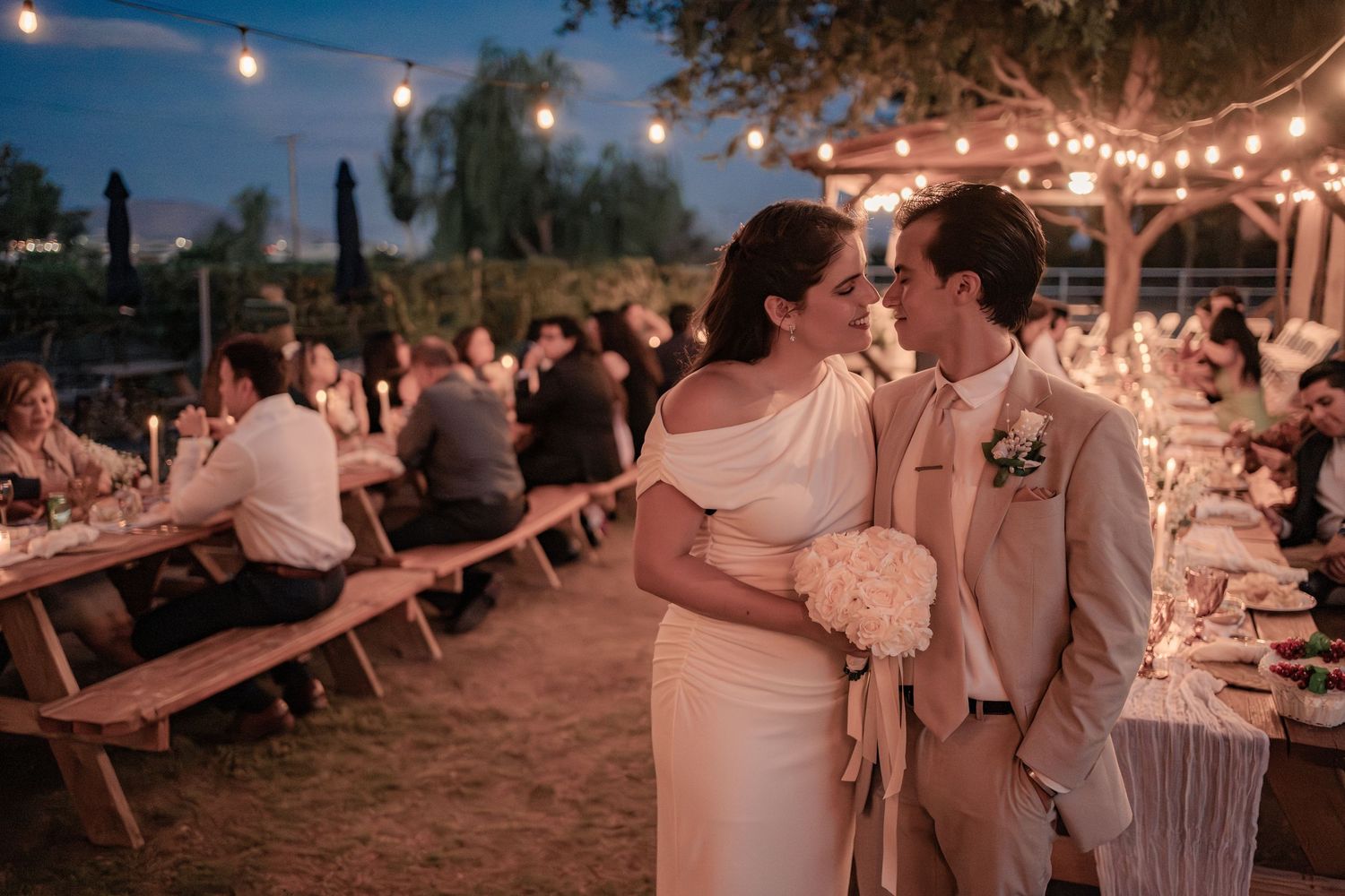An elegant outdoor evening wedding reception with string lights and long wooden tables in a desert setting at dusk.