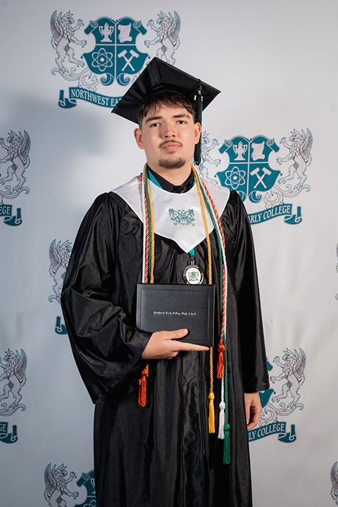Graduate in black cap and gown poses with diploma in front of university backdrop with heraldic emblems.
