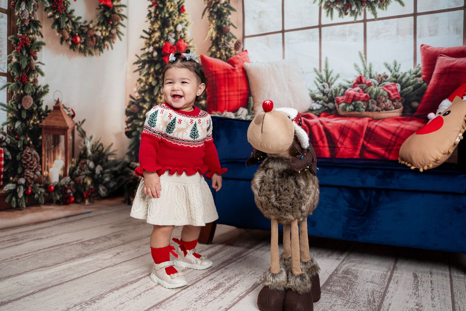 A child in a red holiday dress sits on a wooden floor surrounded by festive Christmas decorations.