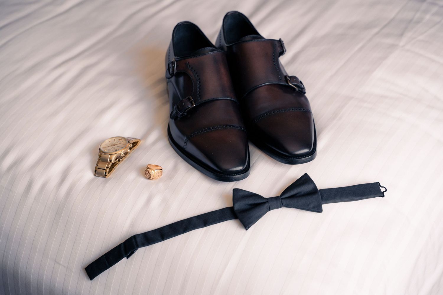 Black dress shoes and formal accessories laid out on white bedding in preparation for a special event.