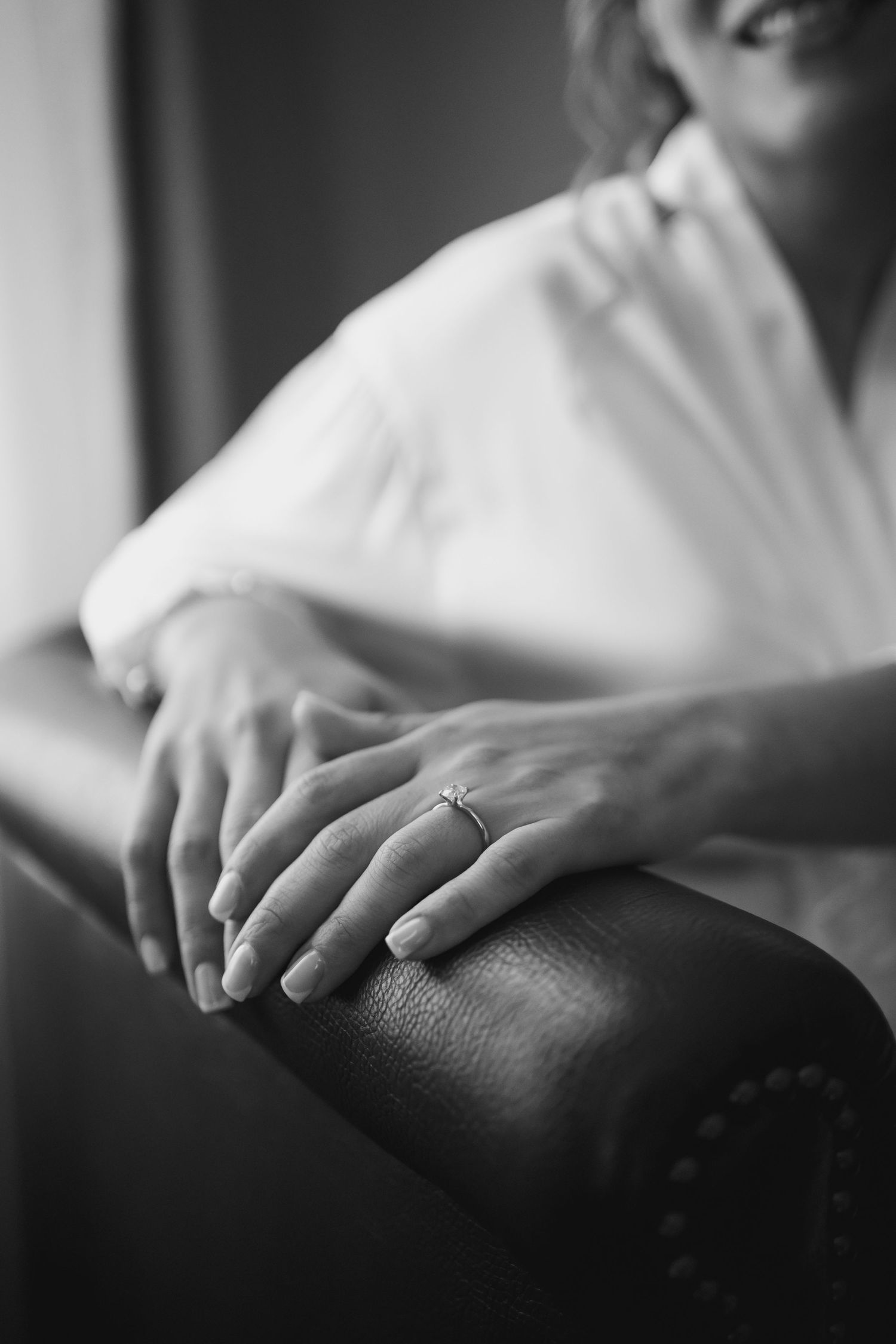 Black and white photo of a bride displaying her diamond engagement ring while resting hands on leather chair.