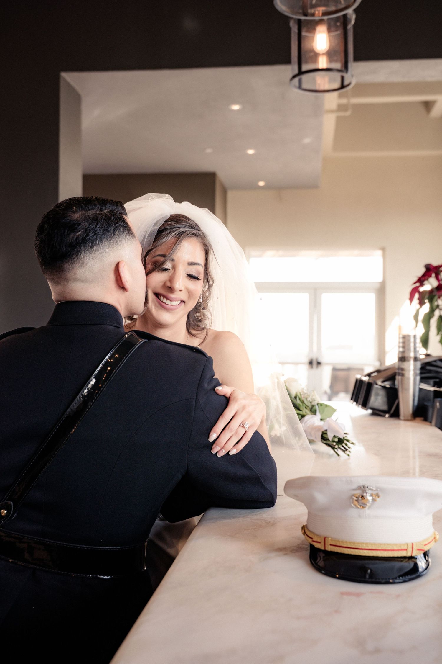 Smiling bride in veil embraces uniformed military groom, with white officer cap on marble counter in elegant venue.