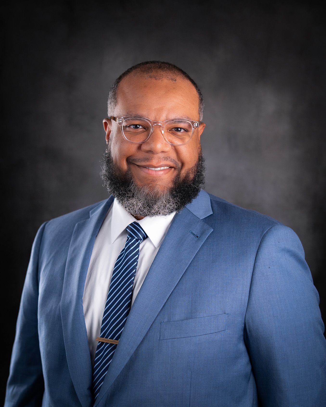 Professional headshot portrait of someone wearing a blue suit, striped tie and glasses against a dark background.