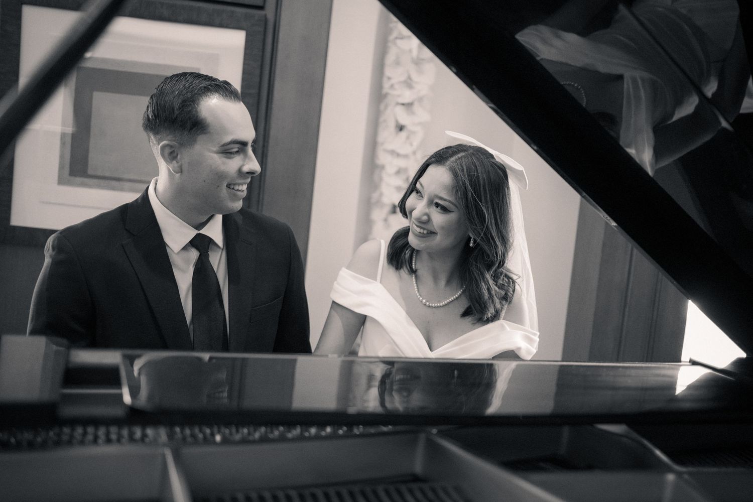 Black and white photo of a couple smiling at each other beside a grand piano.