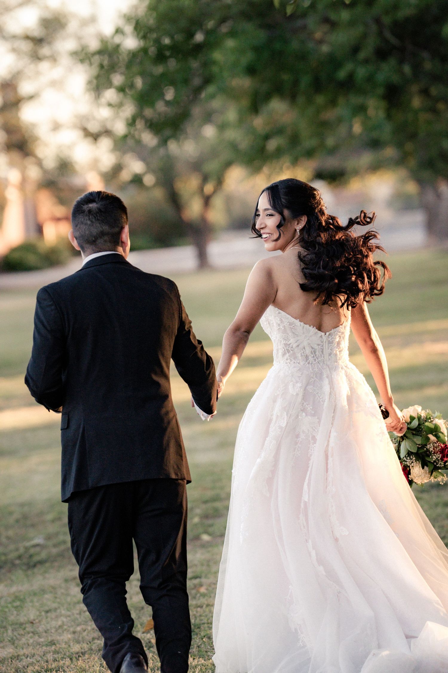 A bride in a white lace gown smiles back while holding hands with her groom in a black suit outdoors.