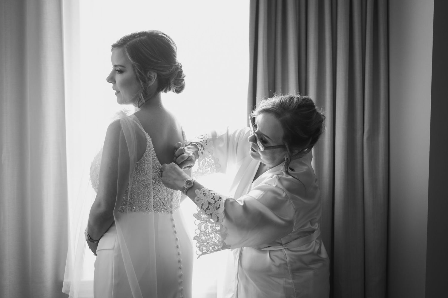 Black and white photograph of wedding dress preparations by window light.