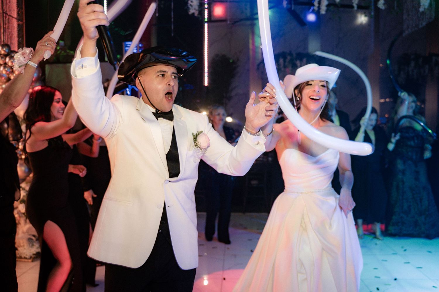 Bride and groom dancing joyfully at wedding reception, holding light-up wands while wearing headphones.