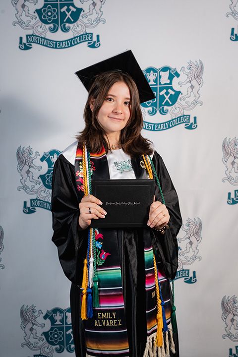 Graduate in black cap and gown holds diploma while posing in front of decorative backdrop with school emblem.