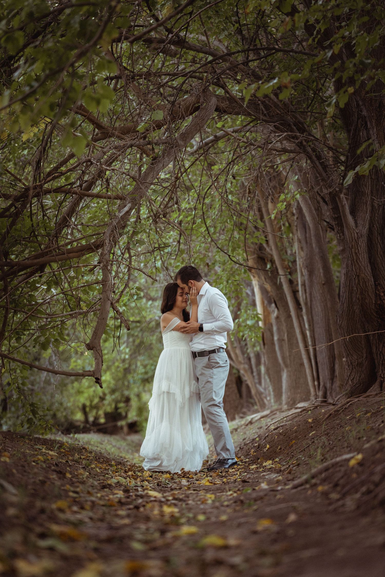 A romantic couple in white attire share an intimate moment along a tree-lined forest path.