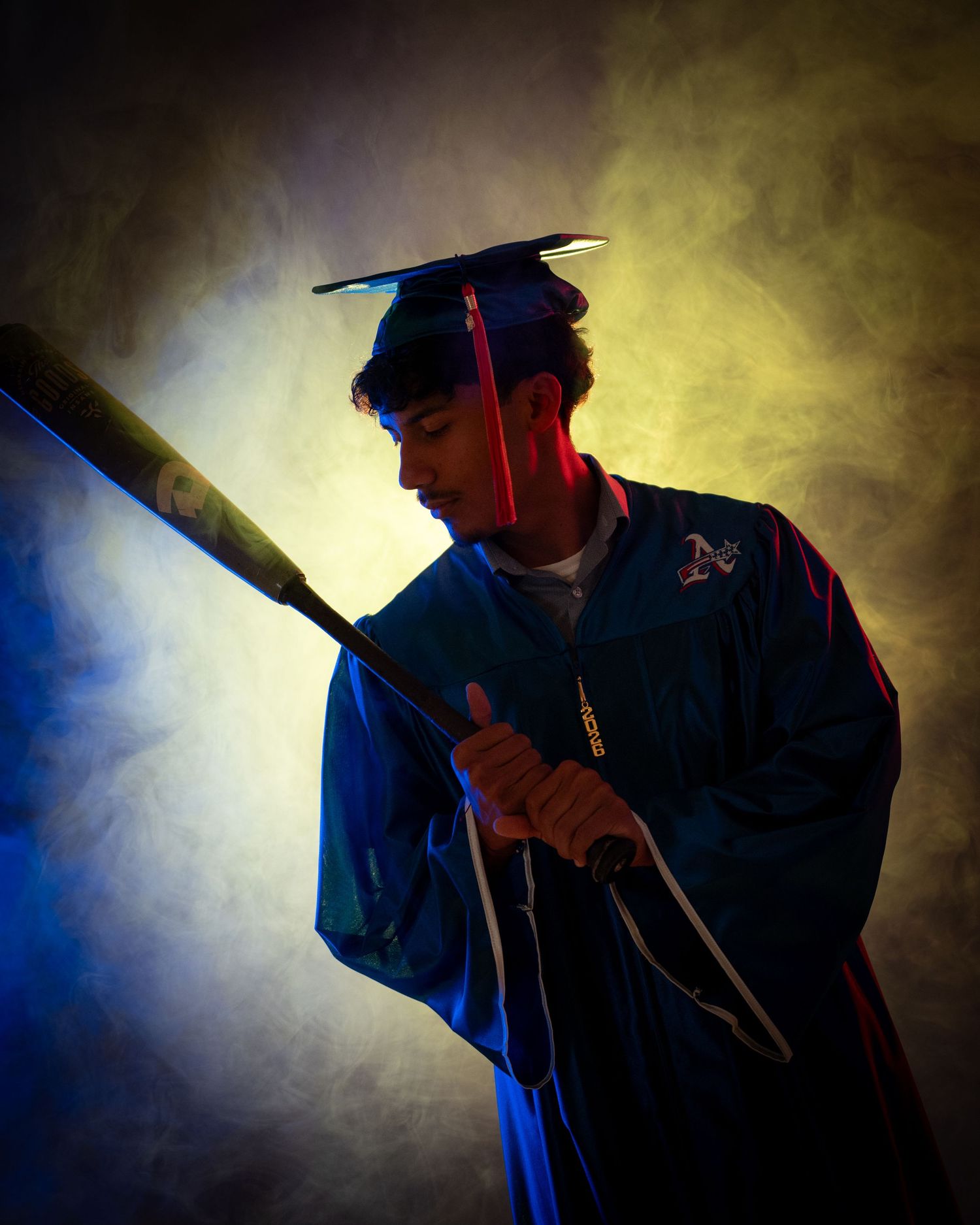 Graduate in blue cap and gown holds baseball bat dramatically under colorful studio lighting with smoke effects.